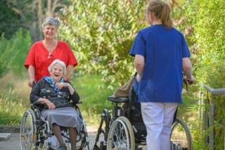 Zwei Altenpflegerinnen schieben jeweils eine Bewohnerin im Rollstuhl und begegnen sich im Garten.