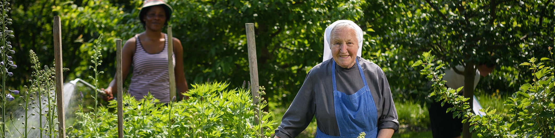 Eine Oberzeller Franziskanerin steht im Klostergarten und gießt mit einer Gießkanne und lächelt zufrieden. Im Hintergrund sieht man eine junge Frau, die mithilft.