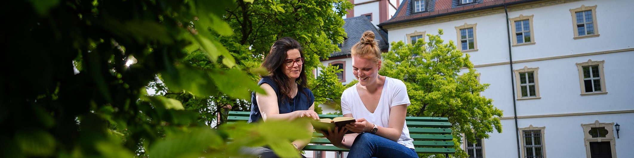 Eine Oberzeller Schwester sitzt mit einer jungen Frau auf einer Gartenbank vor dem Klostergebäude. Die beiden unterhalten sich im Rahmen des Angebots Kloster auf Zeit.