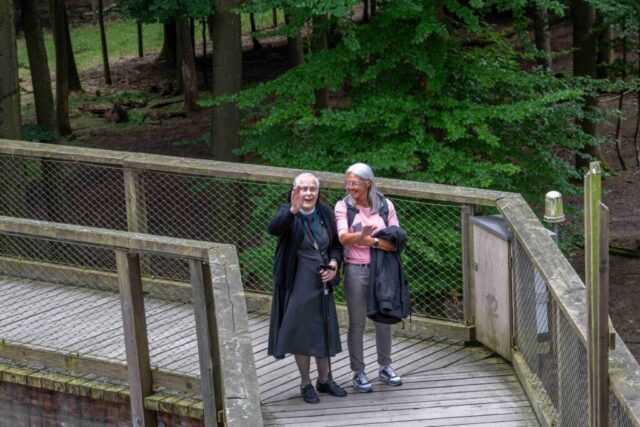 Unterwegs auf dem Holzsteg, eine Schwester winkt in die Kamera. Sie wird von einer Frau gestützt.