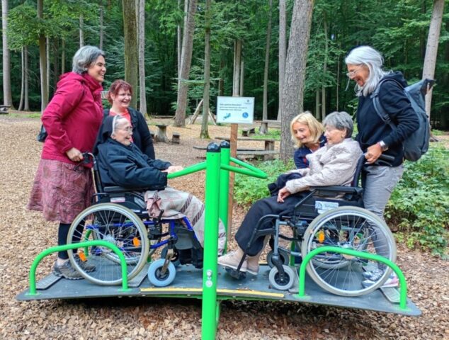 Zwei Rollstuhlfahrerinnen haben Spaß auf der Rollstuhlwippe, die Begleitpersonen auch.