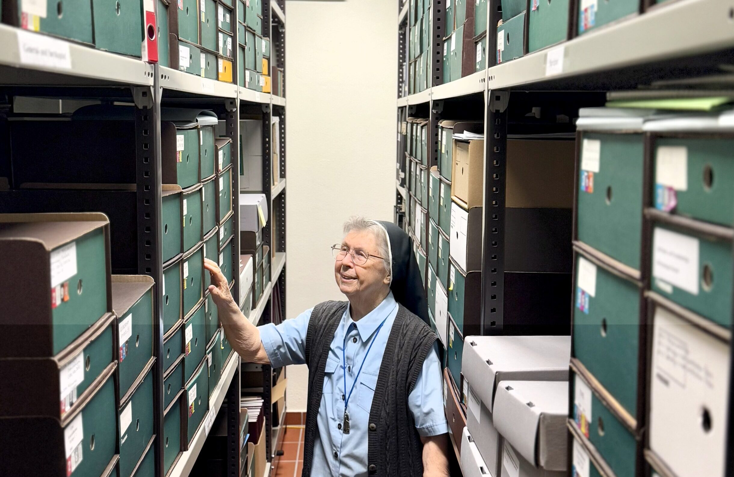 Schwester Erentrud Iselt steht zwischen den Archivregalen, die bis zur Zimmerdecke reichen und in denen viele Pappschachteln gestapelt sind.
