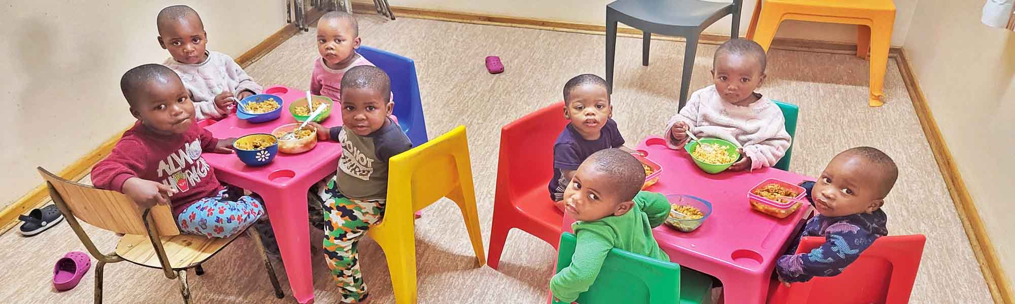 Kleinkinder in Südafrika im Kinderheim St. Joseph sitzen um zwei kleine Tische und essen. Das Essen ist in Südafrika kaum noch zu bezahlen und daher sind die Kinder auf Spenden angewiesen,